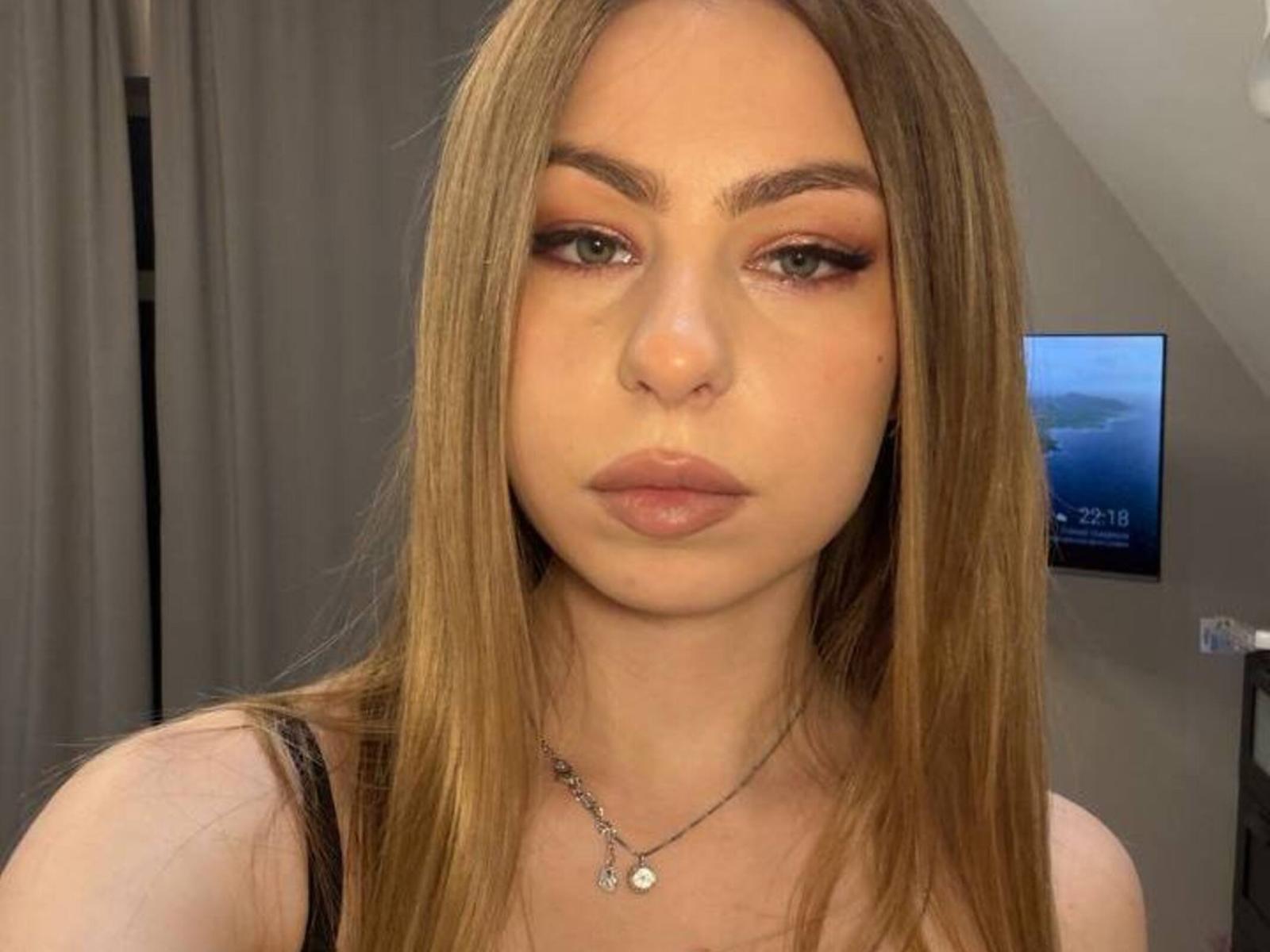 Marinda Boitnott hat langes, glattes, braunes Haar, grüne Augen mit dunklem Eyeliner und volle, rosafarbene Lippen. Sie trägt ein schwarzes Oberteil und eine silberne Halskette mit einem kleinen Anhänger..