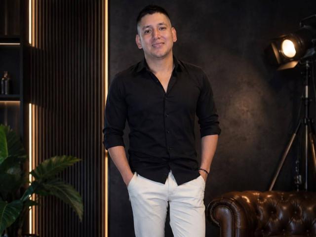 DanteVega, debout, les mains dans des pantalons blancs, une chemise noire à boutons, des cheveux noirs courts, un léger sourire, un canapé en cuir marron sur la droite, un éclairage de studio sur la droite, une plante sur la gauche..