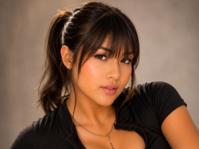 AshlyLee with dark brown hair in a ponytail, wearing a black shirt, gold necklace, and pink lipstick, has a cute, sexy, and attractive face, with a subtle smile.