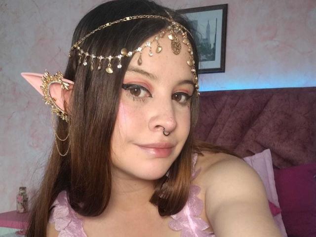 MelaniBecker with pink elf ears, wearing a gold chain headpiece, nose ring, and pink frilled top, looking at the camera with a slight smile. Brown hair, makeup with pink eyeshadow.