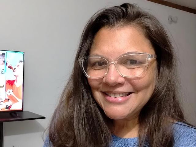 Estela, con i capelli castani, occhiali chiari, sorridente, indossa un maglione blu, mostra i denti e sta in piedi di fronte a una TV con immagini colorate. Il suo naso è piccolo e carino..