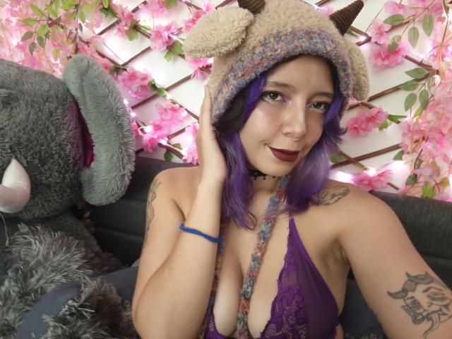 DarckVelvet, con i capelli viola, indossa un cappello morbido, un reggiseno di pizzo viola che lascia intravedere il seno, ha dei tatuaggi, un anello al naso e rossetto scuro, ed è accanto a un enorme coniglio di peluche grigio..