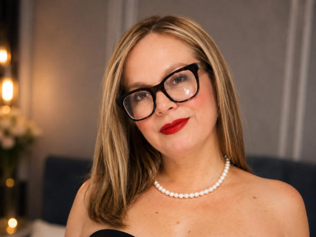 PamelaColin a les cheveux blonds, des lunettes noires, du rouge à lèvres et un collier de perles blanches. Elle sourit, ses épaules sont dénudées, et elle a l'air mignonne et séduisante..