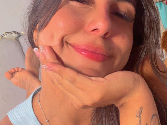 SarahPrincess with a cute smile, pink glossy lips, brown hair, tan skin, hand under chin, visible tattoo on wrist, blue top, and a silver necklace. Her nails are manicured.