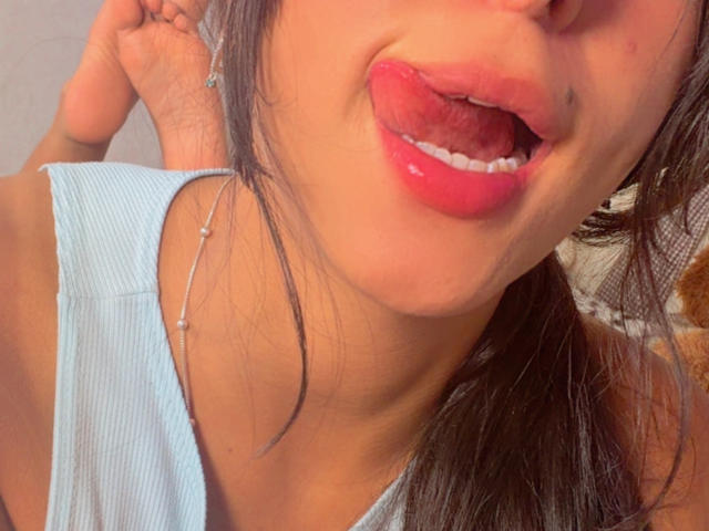 SarahPrincess's face, pink glossy lips, tongue slightly out, blue tank top, dark hair, silver necklace with small beads, holding hair, soft lighting, slight smile, close-up.