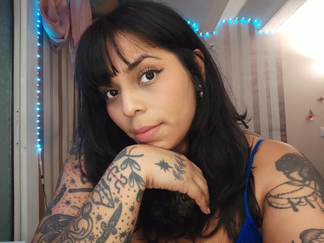 Vrouw met zwart haar, tatoeages op armen en borst, blauwe beha, kin rustend op hand, kijkt naar de camera, blauwe LED-verlichting op de achtergrond, zachte uitdrukking, lichte huid..