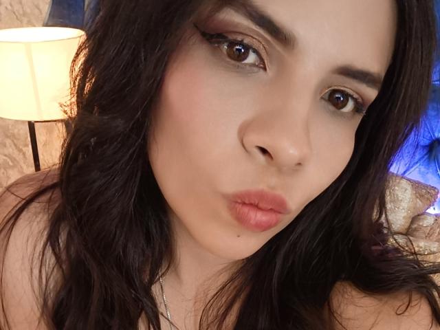 VioletaStone has pouty pink lips, brown eyes with winged eyeliner, and dark wavy hair. She's making a kissy face with a lamp in the background.
