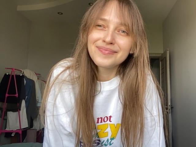 Uma mulher irlandesa chamada Hansche, com cabelos castanhos longos, sorrindo com a língua ligeiramente para fora, vestindo uma camisa branca, em pé em um quarto, com uma expressão fofa e inocente..