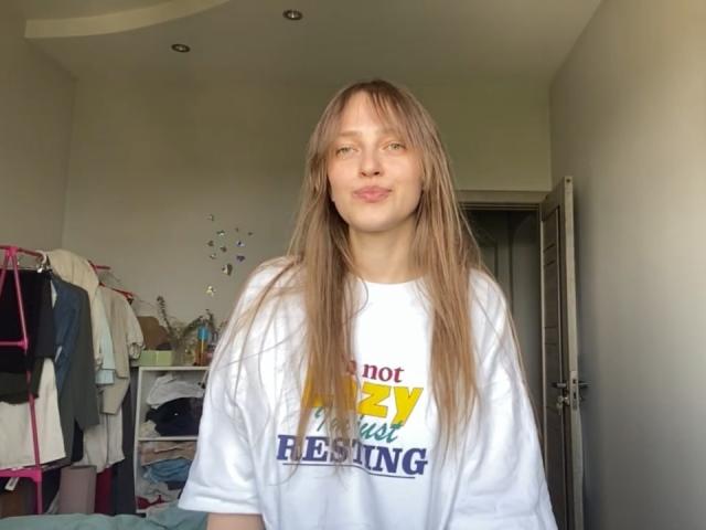 IrishHansche with long brown hair, pouty lips, wearing a white shirt, standing in a room, looking at the camera, hands by sides, no visible boobs, pussy, or ass.