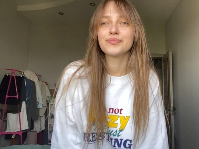 Ein Mann mit irischen Wurzeln, mit langem, glattem, braunem Haar, einem weißen Sweatshirt und einem leichten Lächeln, steht in einem Raum, im Hintergrund sind Kleider an rosa Kleiderbügeln zu sehen..