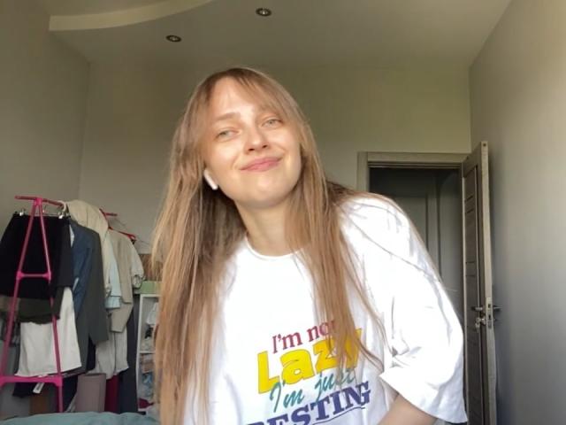 Een IerseHansche met lang blond haar, een wit shirt, die lichtjes glimlacht, met een lichte huid, een kleine neus, draagt witte oordopjes en staat in een kamer met kleding aan een roze hanger..