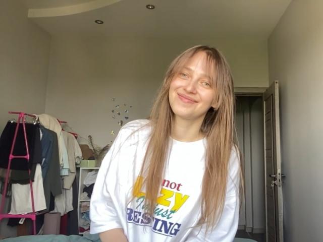 Ierse Hansche met lang blond haar, lachend, gekleed in een witte blouse, staand in een kamer met kleding aan een roze hanger en een open deur..