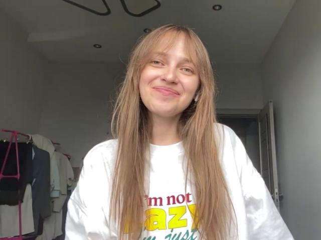 IrishHansche has long, straight brown hair, smiling with pink lips, wearing a white shirt, and has a slim, cute face. Her tits are small, and she's standing in a room.