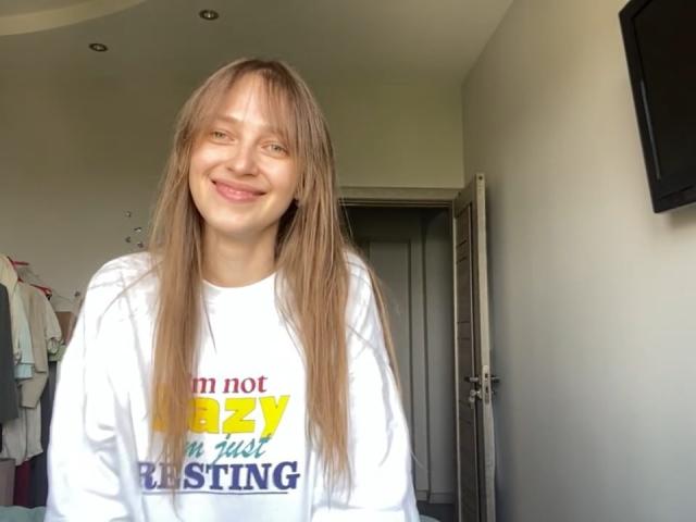 Uma mulher irlandesa, com longos cabelos loiros e lisos, sorrindo, vestindo uma camisa branca de manga comprida, em pé em um quarto com uma televisão à direita e uma porta aberta atrás..