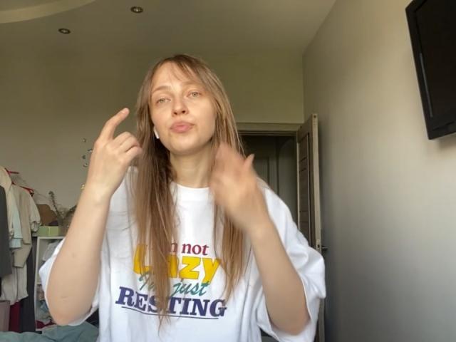 IrishHansche with long blonde hair, white oversized shirt, and puffy lips, making a "rock on" hand gesture, looking slightly annoyed.