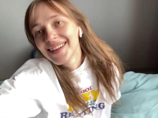 Een Ierse Hansche met lang bruin haar, lachend, gekleed in een wit shirt, met witte oordopjes, een lichtblauwe kussen en een gele vogelprint op het shirt..