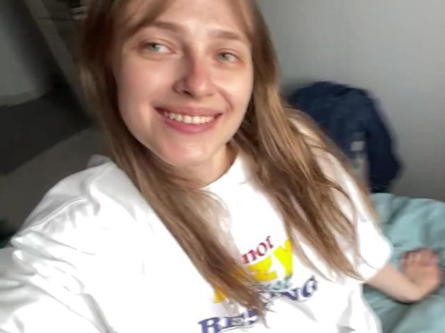 IrishHansche, met haar lichtbruine haar, witte shirt en roze lippen, heeft een schattig gezicht en kleine borsten. Ze maakt een selfie en heeft een ontspannen en vrolijke uitdrukking..