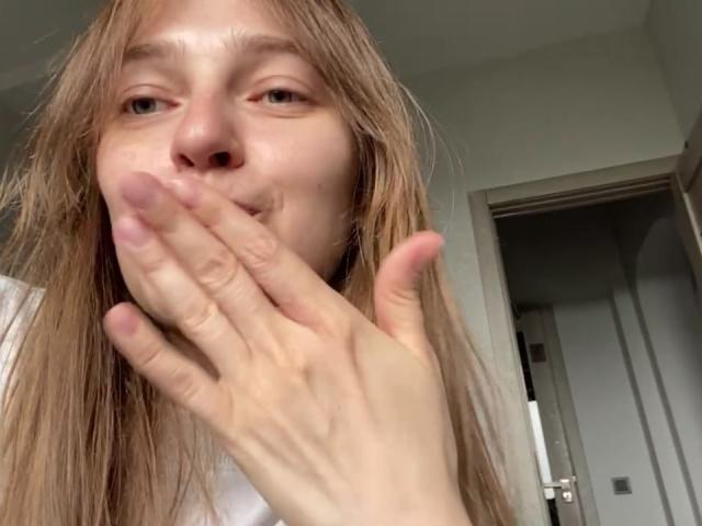 Een Ierse vrouw met blond haar, die haar mond bedekt met een hand, waarbij haar neus en groene ogen zichtbaar zijn. Ze heeft een lichte huid en draagt een witte blouse..