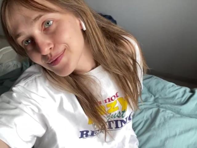 IrishHansche with light brown hair, green eyes, and a slight smile, wearing a white shirt, sitting on a blue bed, wearing white earbuds, looking up at the camera.