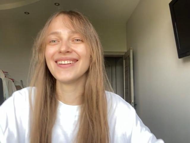 Een IerseHansche met lang, steil blond haar, lachend, gekleed in een wit shirt, staand in een kamer, lachend met zichtbare tanden, haar is iets rommelig, kleine neus, lichte huid..