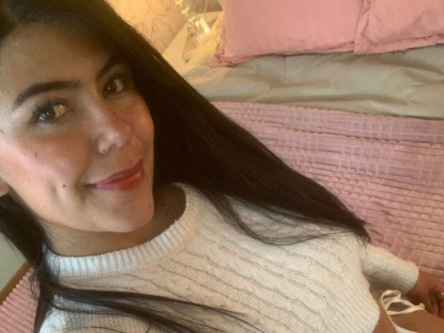 CatalinaReiyna with long black hair, brown eyes, and red lipstick, wearing a white ribbed sweater, smiling at the camera, showing a small mole on her right cheek.