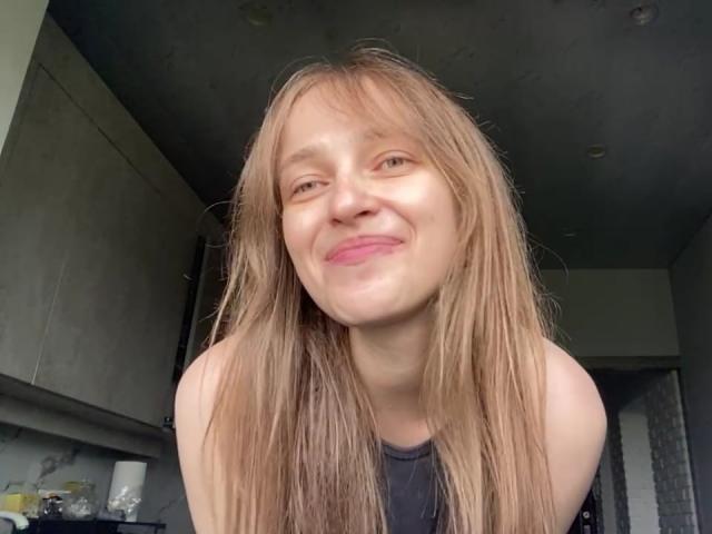 Een Ierse vrouw met lang, vuilblond haar, die glimlacht met roze lippen, gekleed in een zwart topje zonder mouwen, waarbij haar blote schouders te zien zijn, in een moderne keuken..