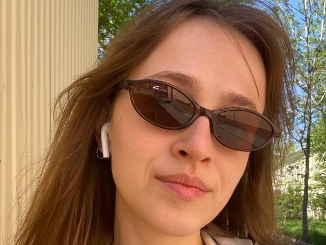 Una mujer irlandesa, con ojos marrones, usando gafas de sol ovaladas negras, auriculares blancos y cabello castaño, sonriendo ligeramente, sin maquillaje, con piel natural, de pie afuera, en un día soleado..