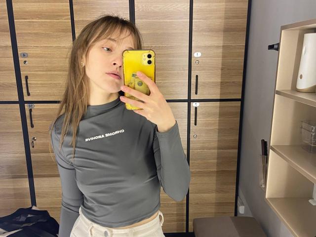 Irische Frau mit mittelgroßer Brust, graues Langarmshirt, hält ein gelbes Telefon, Spiegel-Selfie, Hintergrund mit Holztüren, hellbraunes Haar, weiße Hose, lässiger Look, leichtes Schmollen im Gesicht..