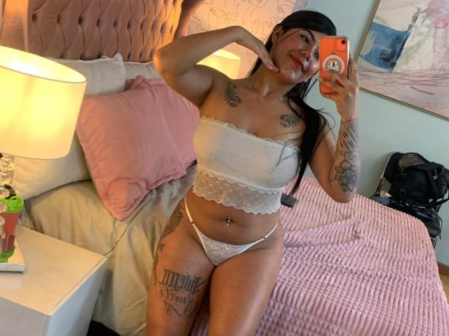 CatalinaReiyna with tattoos, wearing white lace top and thong, sitting on pink bed, taking selfie with orange phone, showing her tits and pussy. She's smiling, touching her chin.