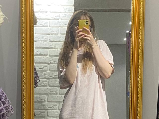 Una ragazza di origine irlandese, con una maglietta rosa larga, lunghi capelli castani, che tiene in mano un telefono giallo, un selfie allo specchio, uno specchio con cornice dorata, un muro di mattoni bianchi, aspetto leggermente trasandato..