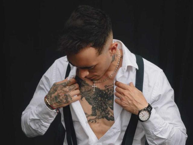 JasonKiing met tatoeages, een wit shirt, zwarte braces, een kruisbeeldketting en een horloge, kijkt naar beneden, handen op de borst, kort donker haar, zwarte achtergrond..