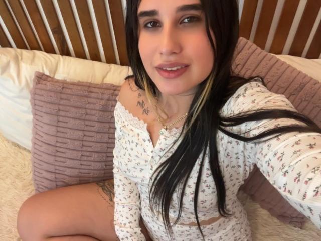 KatieDesirX, con cabello negro largo, vestida con una blusa blanca con estampado floral, sentada en una cama con almohadas rosas, mostrando sus senos y un tatuaje en su muslo derecho. Sonriendo, mirando a la cámara..