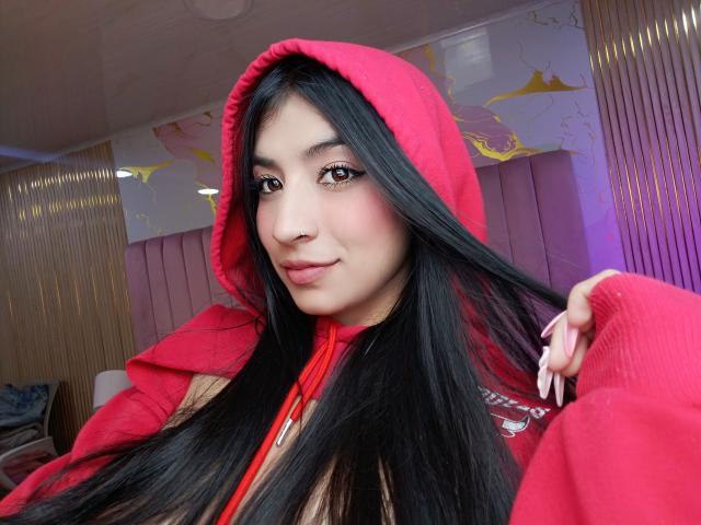 Naia Sanders, avec de longs cheveux noirs, portant un sweat à capuche rouge avec la capuche relevée, tenant ses cheveux avec sa main droite, des ongles roses, souriant légèrement, des yeux marrons, une peau claire, dans un environnement intérieur confortable..