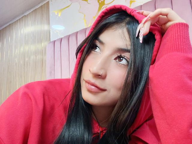 NaiaSanders with long black hair, red hoodie, and pink nails, has a nose ring and brown eyes, looking up with a slight smile. Her tits are not visible.