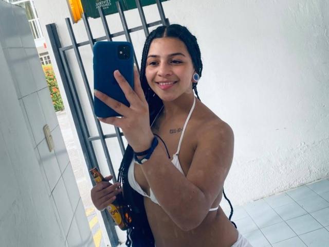 LonxEva with long black braids, smiling, wearing a white bikini top, holding a blue phone, showing a tattoo on her chest, standing in front of a white wall.