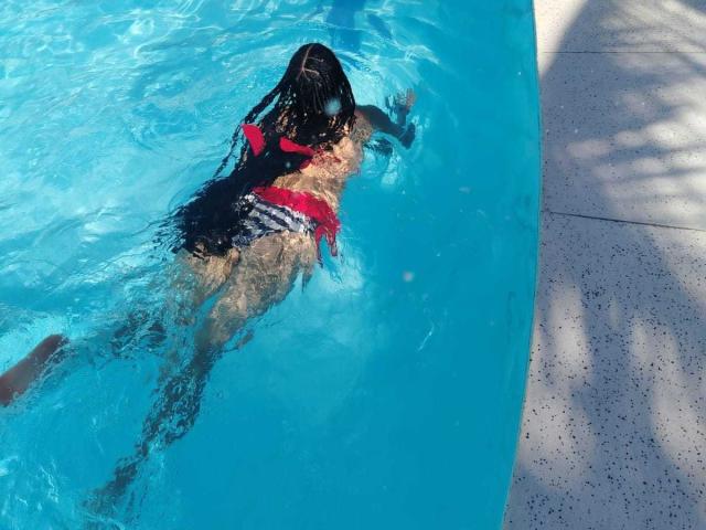 LonxEva, black-haired, wearing a red and black bikini, laying on her stomach in a blue pool, with her ass and back arched, water rippling around her.