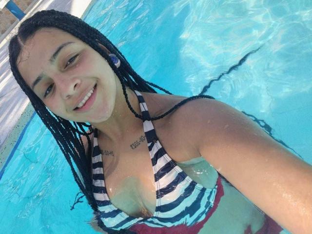 LonxEva with wet braids, smiling in a blue pool, wearing a striped black and white bikini top, showing her small tits and red bottoms, holding a selfie.