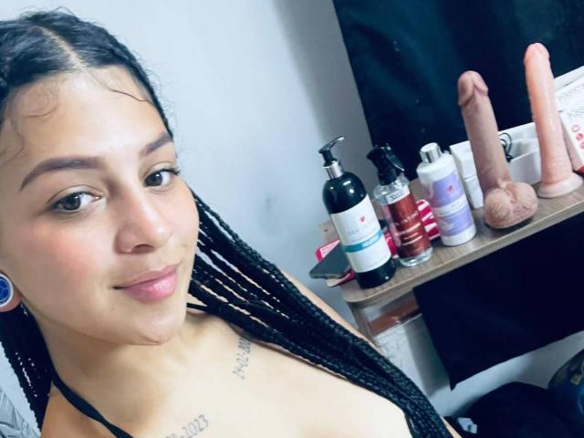 LonxEva with braided black hair, light brown skin, small tattoos, blue earrings, smiling at camera, showing cleavage, with a shelf of dildos and sex toys in the background.