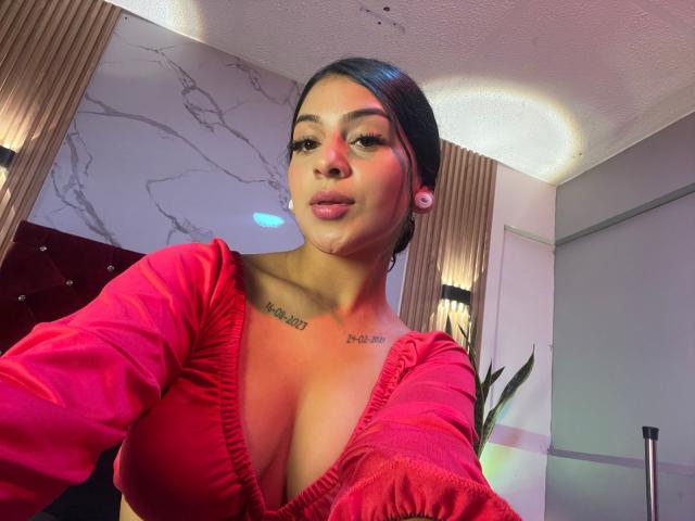 LonxEva, avec ses cheveux foncés, sa robe rose et ses seins volumineux, ses lèvres gonflées, des tatouages sur la poitrine, des écouteurs roses, dans une pièce avec des murs en marbre et des panneaux en bois..