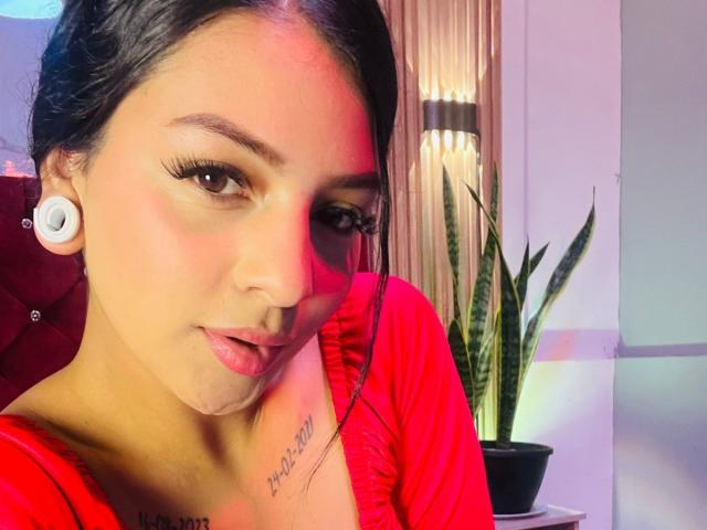 LonxEva, com cabelo escuro, blusa vermelha e fones de ouvido brancos, exibindo seus seios e uma tatuagem discreta, olhando para a câmera com cílios longos e um leve sorriso..