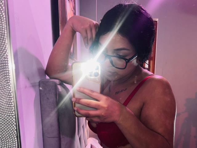 LonxEva with short black hair, glasses, and red bra, taking a mirror selfie with a flash, showing her small tits and tattoo above her right breast.