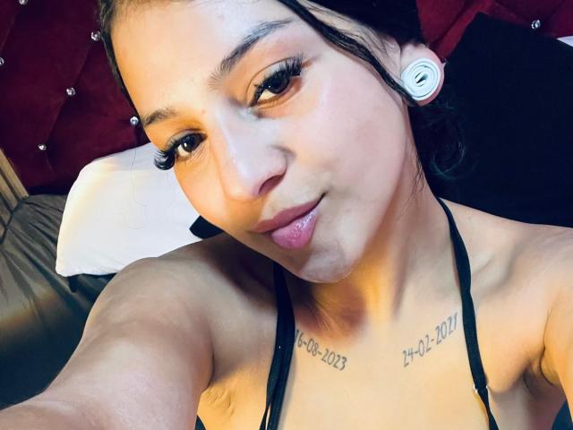 LonxEva tiene la piel de color marrón claro, pechos pequeños, tatuajes en el pecho, delineador de ojos negro, y lleva auriculares blancos, sonriendo a la cámara con un labio rosa ligeramente entreabierto..