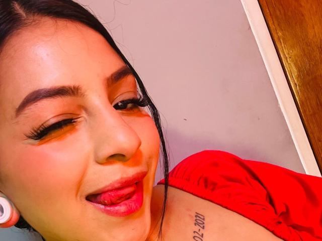 LonxEva, con la pelle abbronzata, i capelli neri e il rossetto rosso, che fa la linguaccia, indossa una maglietta rossa, ha un tatuaggio sulla spalla e degli auricolari bianchi, appare giocosa con l'eyeliner..