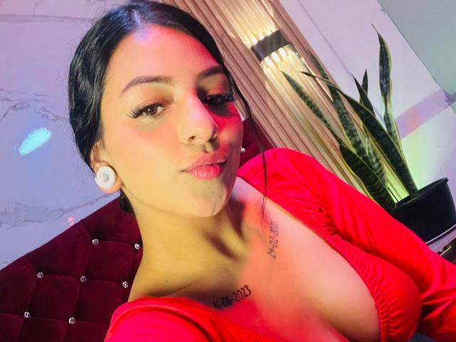 LonxEva with medium brown skin, black hair, red top, showing her tits, white earbuds, tattoo above her cleavage, and a slight smile. Background includes a red tufted headboard.