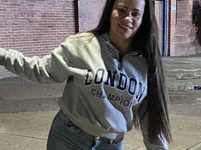 CharllotI, con cabello castaño largo, sudadera gris con la inscripción "London Champions" y jeans azules, sonriendo, inclinándose hacia adelante, con el brazo derecho levantado, fondo de calle, noche, informal, segura de sí misma, atractiva..