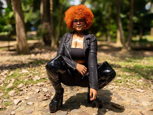 Alice Jelanni mit orangefarbener Afrikakopfbedeckung, einer schwarzen Lederjacke, einem schwarzen BH und glänzenden schwarzen Latex-Hosen, hockt auf einem steinernen Pfad, trägt Plateauschuhe, in einem sonnendurchfluteten Park..