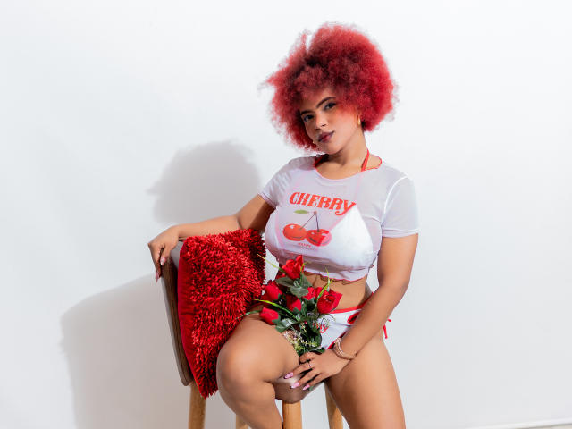 AliceJelanni mit roter Afrofrisur, weißem, durchsichtigen Oberteil und rotem Kirschmuster, sitzt auf einem Stuhl mit einem roten, flauschigen Kissen, hält Rosen, trägt rote Unterwäsche und zeigt ihre Brüste und ihre Vulva..