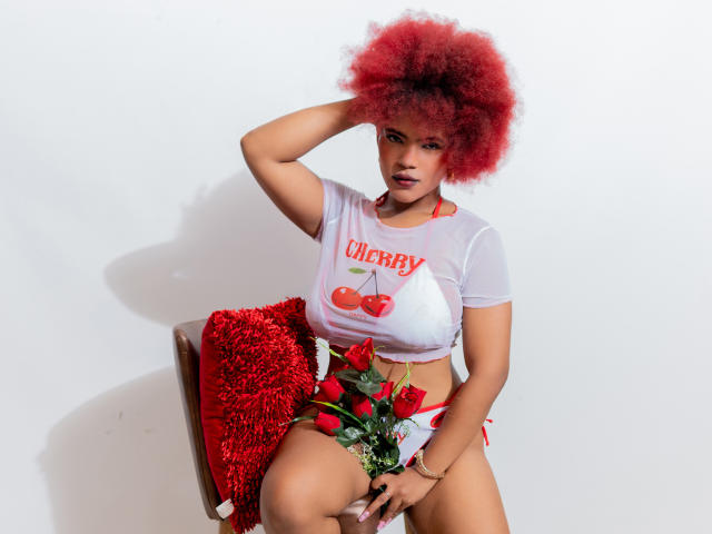 Alice Jelanni mit roter Afrofrisur, trägt ein weißes, durchsichtiges Oberteil, rote Kirschen und weiße Shorts, sitzt auf einem Stuhl mit einer roten, flauschigen Decke, hält rote Rosen und schaut in die Kamera..
