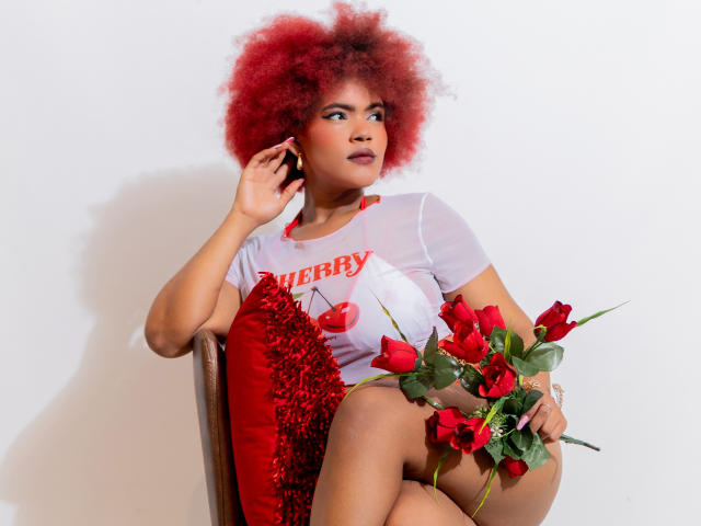 AliceJelanni mit rotem Afro, trägt ein durchsichtiges weißes Oberteil, hält rote Rosen, sitzt auf einem Stuhl mit einem roten Kissen, telefoniert, blickt zur Seite, kurvige Figur, süß, sexy..