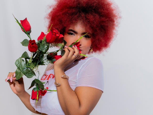 Alice Jelanni mit rotem Afro, die Rosen hält, ein durchsichtiges weißes Oberteil trägt, goldene Ohrringe und Armbänder, eine Rose beißt, rosa Nägel, weißer Hintergrund, ihre Brüste sind durch das Oberteil sichtbar..